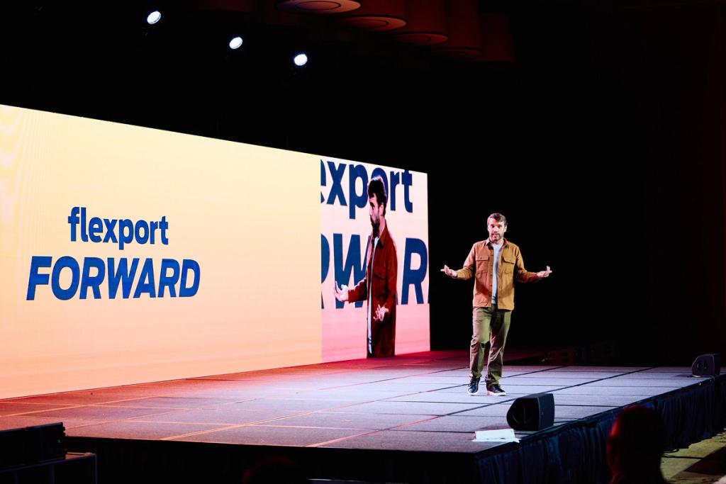 Speaker gestures on stage before a large "flexport FORWARD" screen at a darkened event venue.