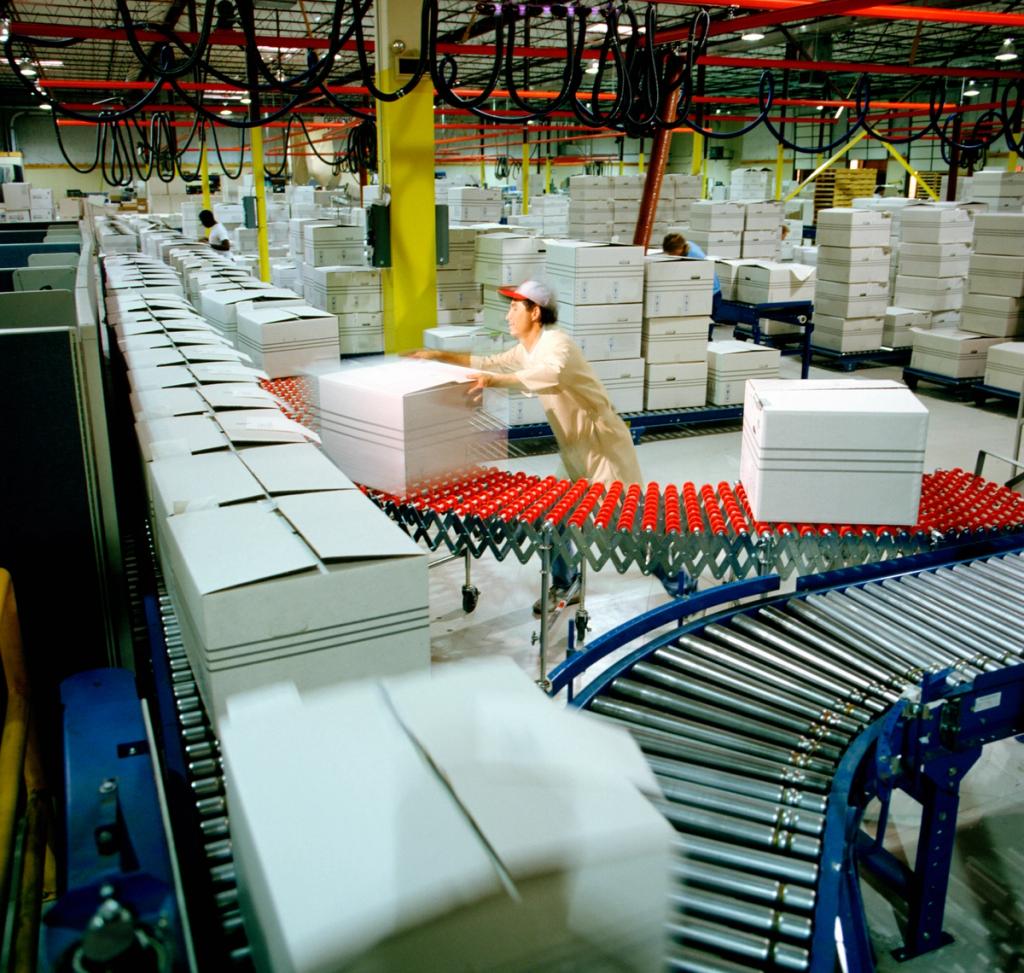 Worker loading flat white boxes onto roller conveyors inside a busy manufacturing and distribution warehouse