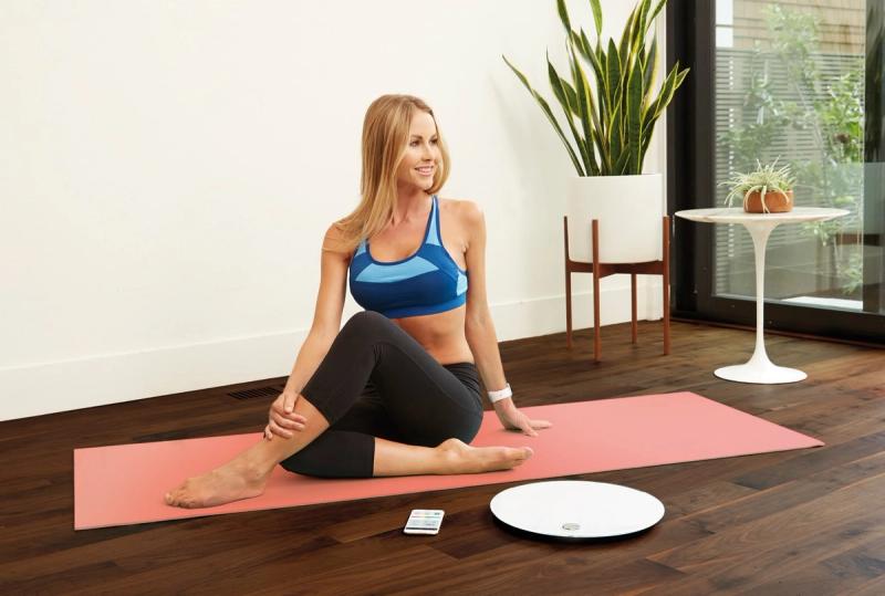 Smiling woman in blue sports bra sits on a pink yoga mat beside a smart scale and phone
