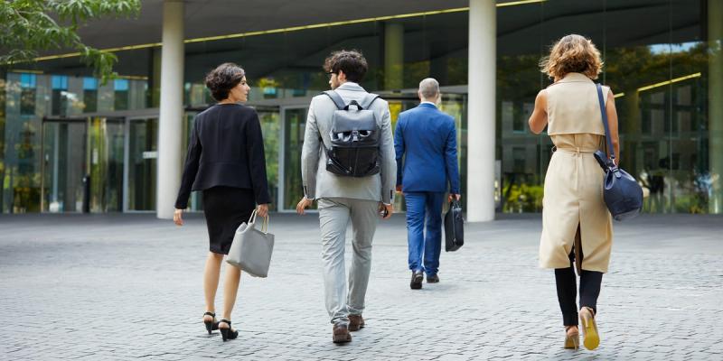 Four business people in suits walk away with bags toward a glass office building entrance
