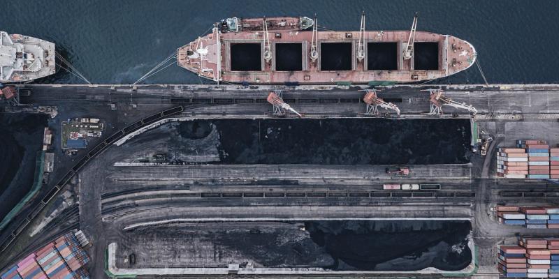 Aerial view of a bulk carrier docked at a coal terminal beside black coal piles and stacked containers