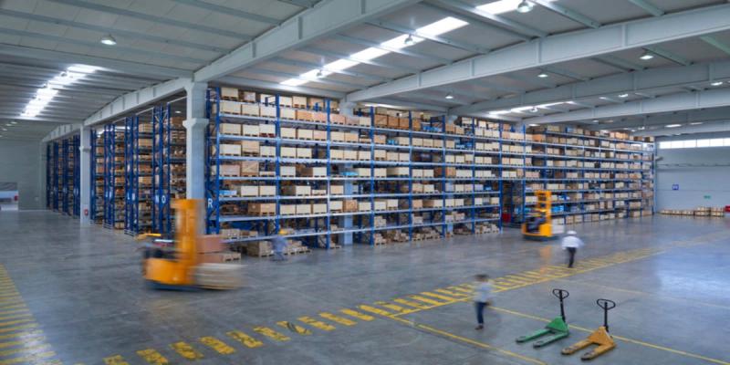 Two workers operating forklifts in a large warehouse with tall blue racks of stacked boxes