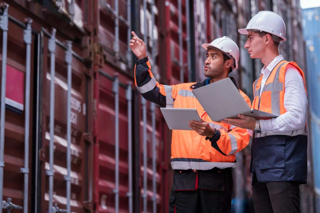 Two customs and compliance team members in hard hats and high-visibility vests reviewing shipment data on a laptop and tablet in a shipping container yard