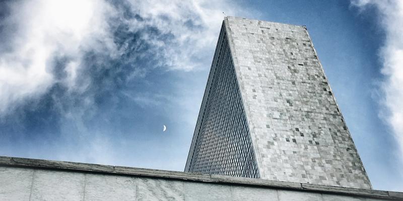 Tall tapered office tower viewed from below against a partly cloudy sky with a crescent moon