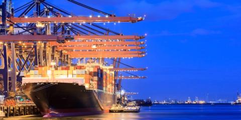 Container ship docked at illuminated port cranes at dusk with city lights on the horizon