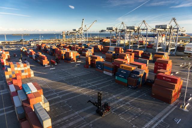 Port container yard with stacked containers, gantry cranes, and a reach stacker beside the water at sunset