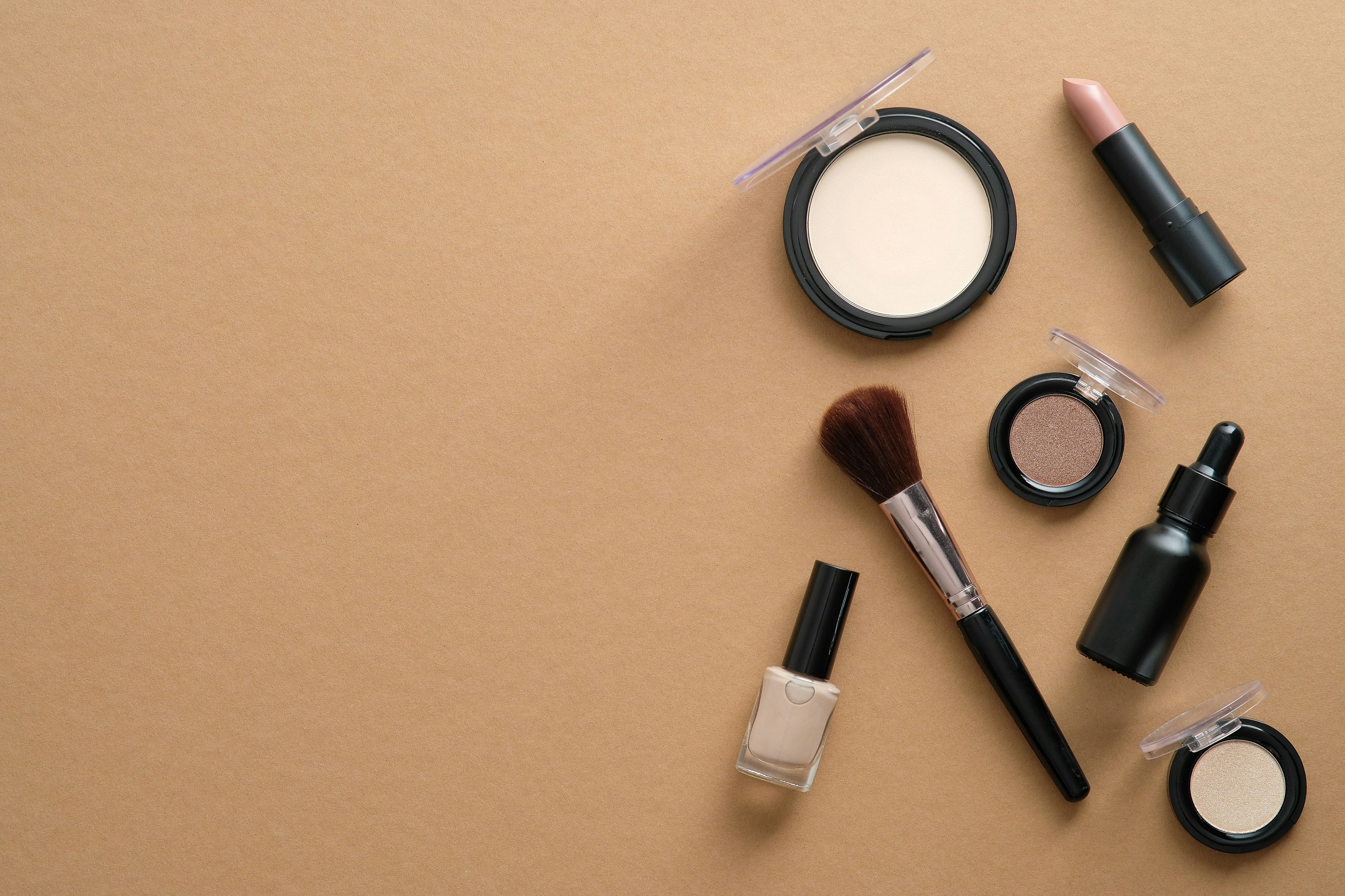 Flat lay of makeup products including eyeshadow, lipstick, nail polish, brush, and skincare serum arranged on a neutral surface.