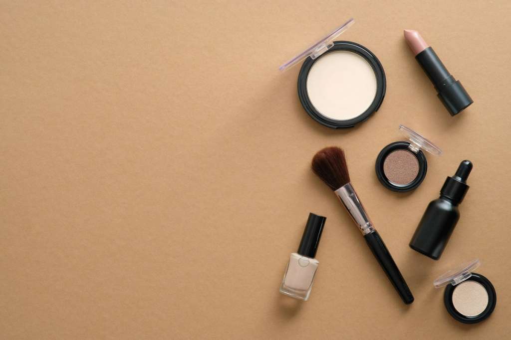Flat lay of makeup products including eyeshadow, lipstick, nail polish, brush, and skincare serum arranged on a neutral surface.