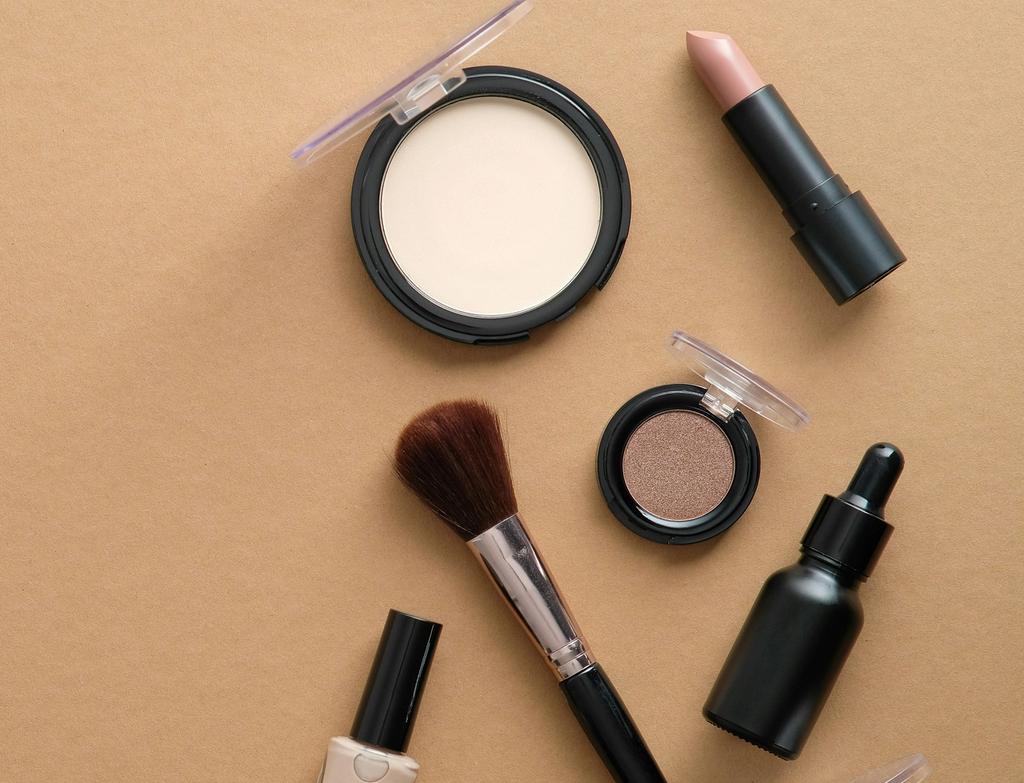 Flat lay of makeup products including eyeshadow, lipstick, nail polish, brush, and skincare serum arranged on a neutral surface.