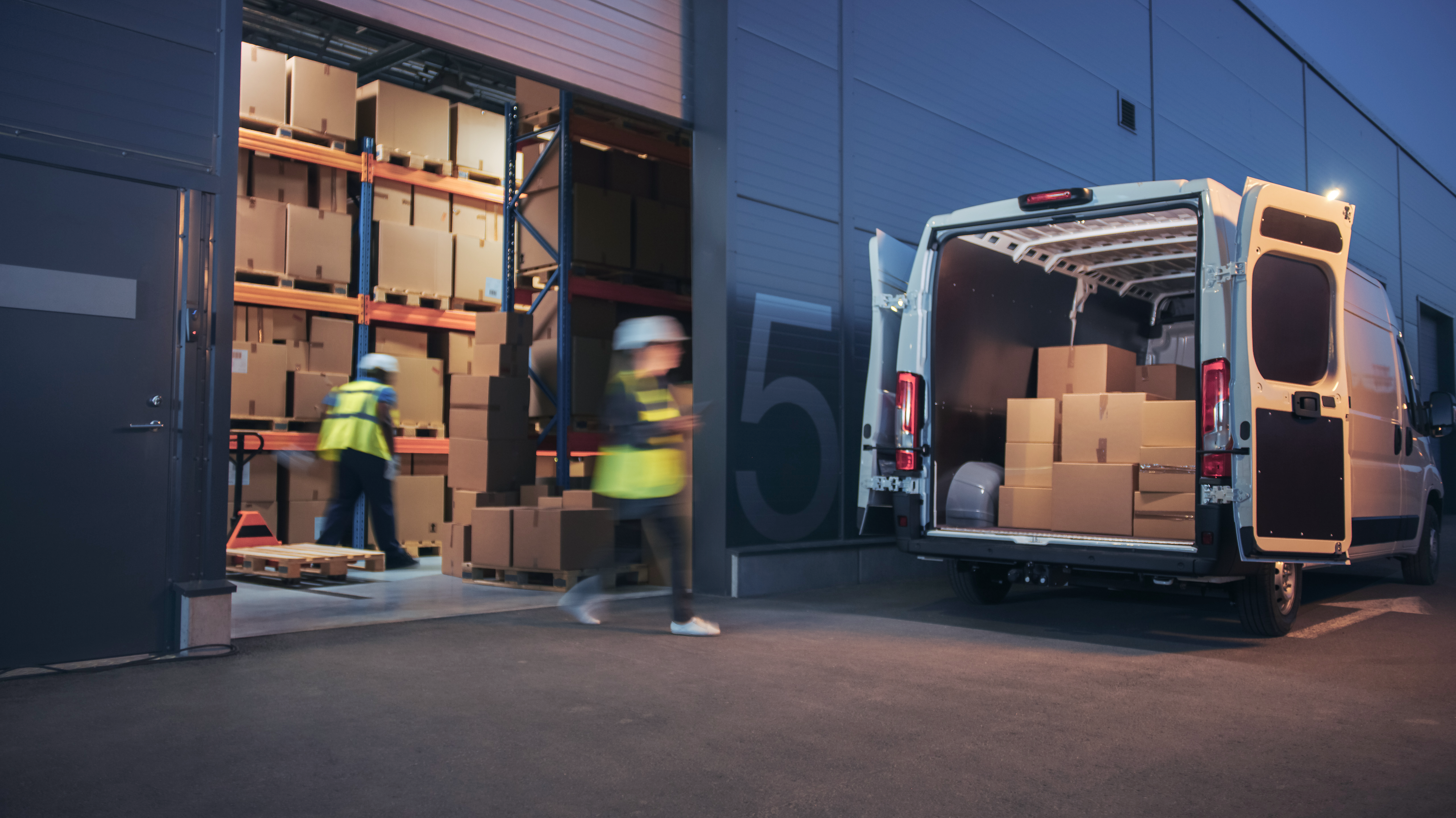 Warehouse workers in safety vests load cardboard boxes into a delivery van.