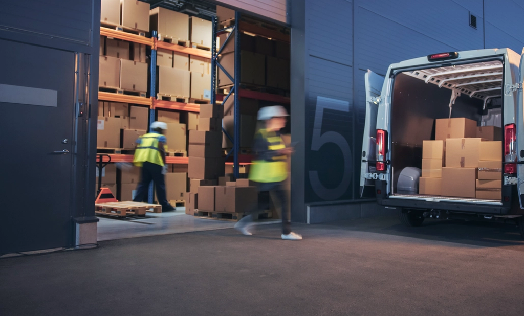 Warehouse workers in safety vests load cardboard boxes into a delivery van.