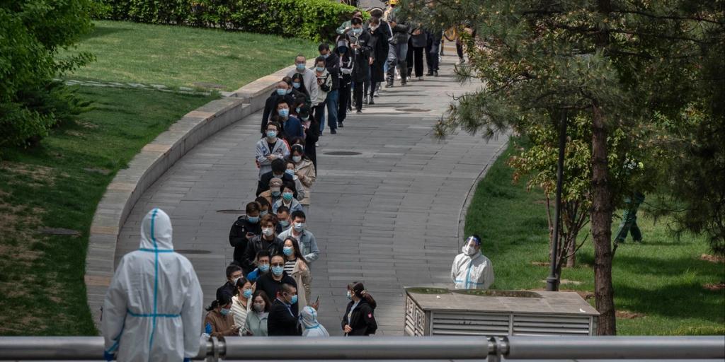 Long line of masked people queuing on a curved park path beside workers in white protective suits