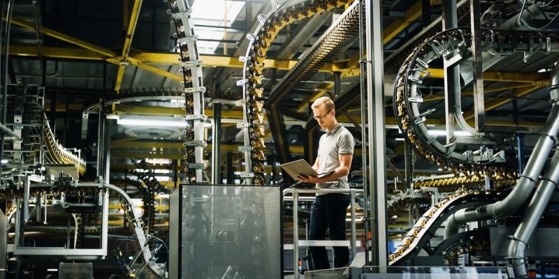 A man with a laptop stands on a platform amid sprawling overhead conveyor systems in an industrial bottling plant.