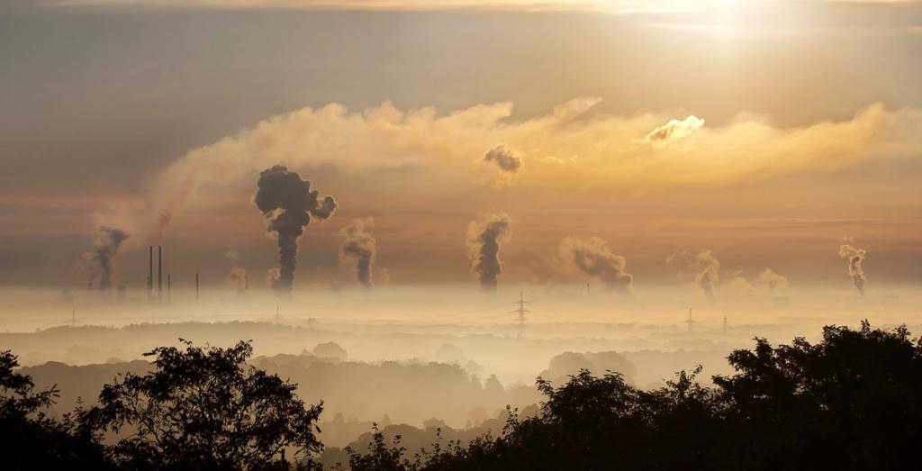 Industrial skyline at sunrise with multiple smokestacks emitting plumes of smoke over a hazy forested horizon