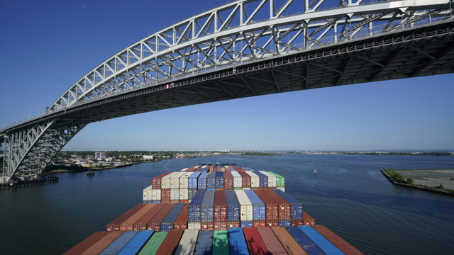 Container ship loaded with stacked shipping containers passing beneath a large steel arch bridge