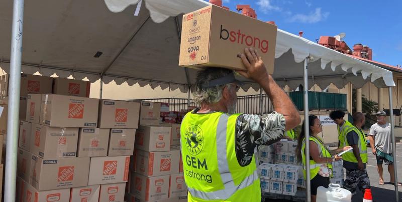 Volunteer in a green GEM bstrong vest carries a relief box past stacks of Home Depot cartons under a tent