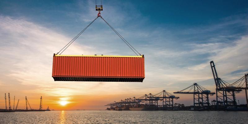 Crane lifts a red shipping container over a harbor at sunset with port gantry cranes in the distance