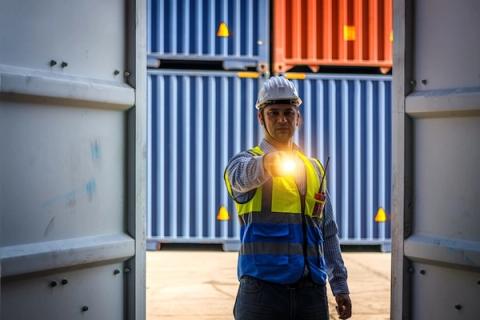 A worker in a white hard hat and hi-vis vest stands in an open container doorway shining a flashlight toward the camera, with blue and orange containers stacked behind.