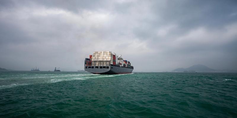 Container ship loaded with stacked containers sailing on green sea under overcast sky, hills in distance