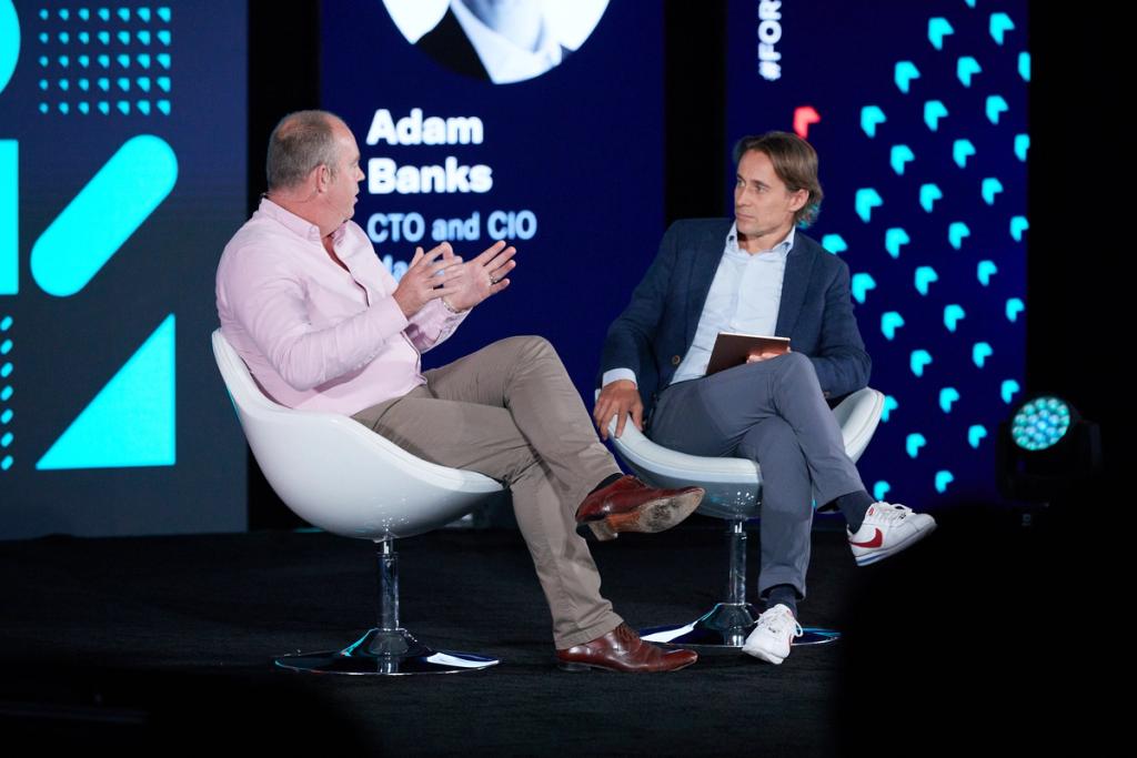 Two men in armchairs on a conference stage in conversation; screen behind reads Adam Banks, CTO and CIO
