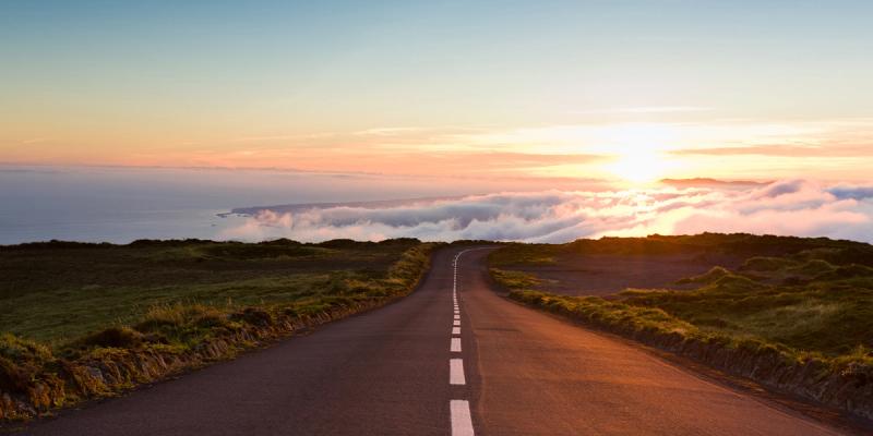 Empty road winding toward a glowing sunset above a cloud-filled valley with grassy verges