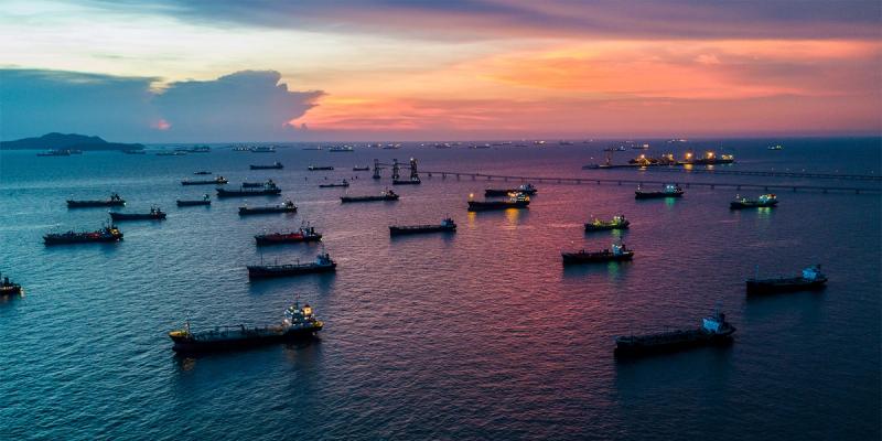 Aerial view of many cargo ships and tankers anchored offshore at sunset under a pink and orange sky