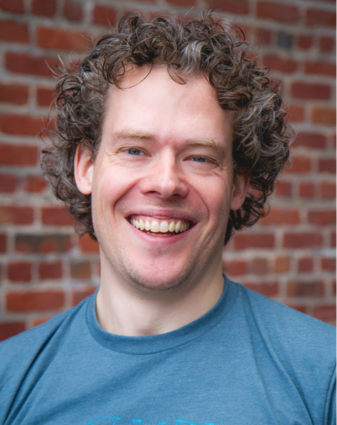 Headshot of a smiling man with curly brown hair in a blue T-shirt against a red brick wall.