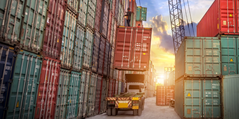 Stacked colorful shipping containers in a port yard with a truck and crane at golden-hour sunset