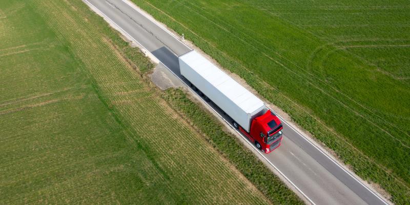 Aerial view of a red semi-truck with white trailer driving a country road between green fields