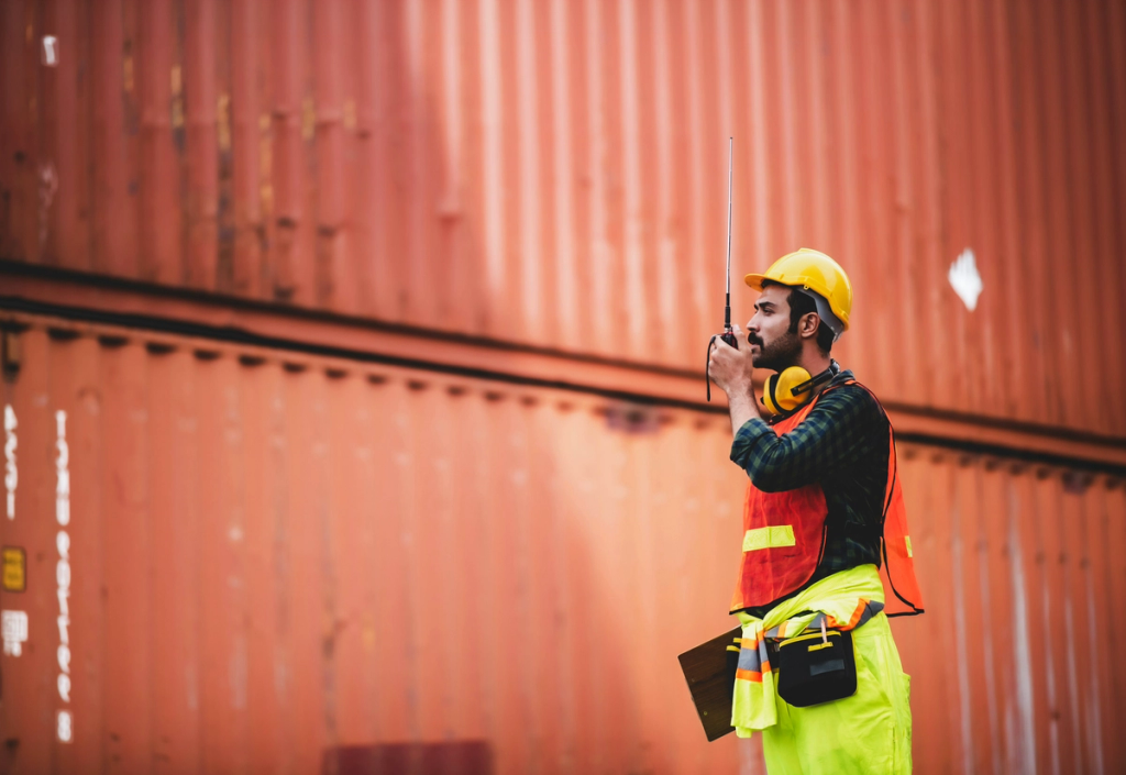 Construction worker wearing safety equipment speaks into a radio while working on containers.