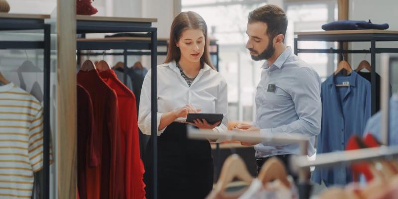 Two retail staff review a tablet between clothing racks in a brightly lit apparel store