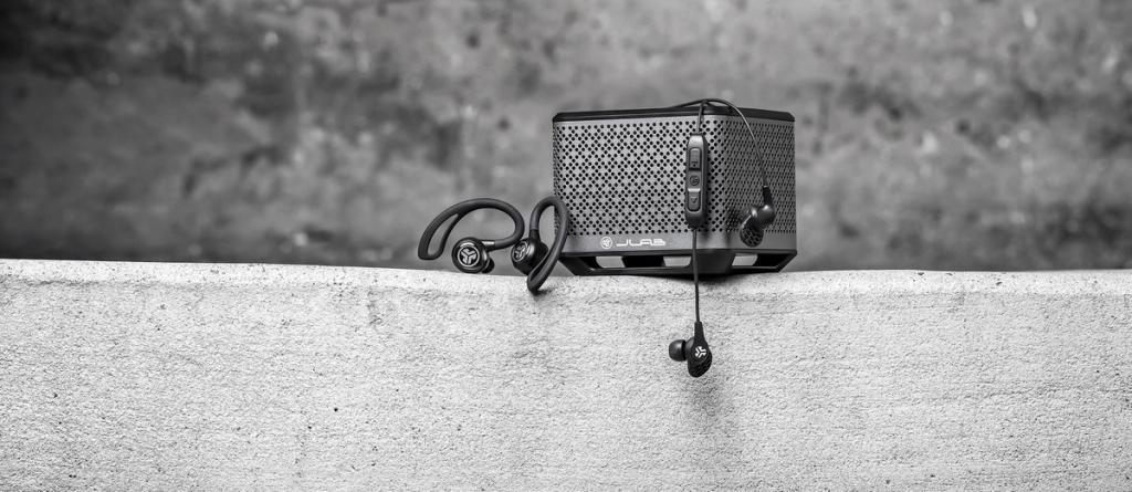 JLab earbuds and a portable Bluetooth speaker on a concrete ledge in black-and-white