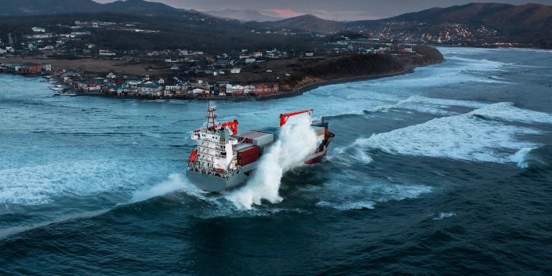 Aerial view of a container ship throwing up spray as it sails past a hilly coastal town