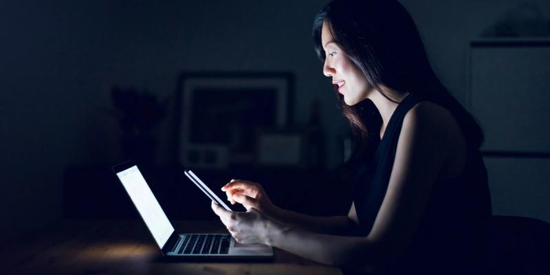 Woman smiling at a laptop screen while holding a smartphone in a dimly lit room at night