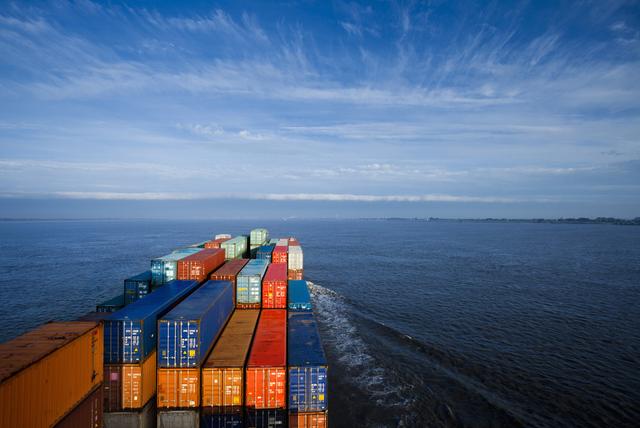 Stacked rows of colorful shipping containers on a container ship's deck sailing across open water under a blue sky