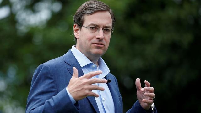 Man in a blue suit and glasses gesturing with both hands while speaking outdoors against a blurred green background