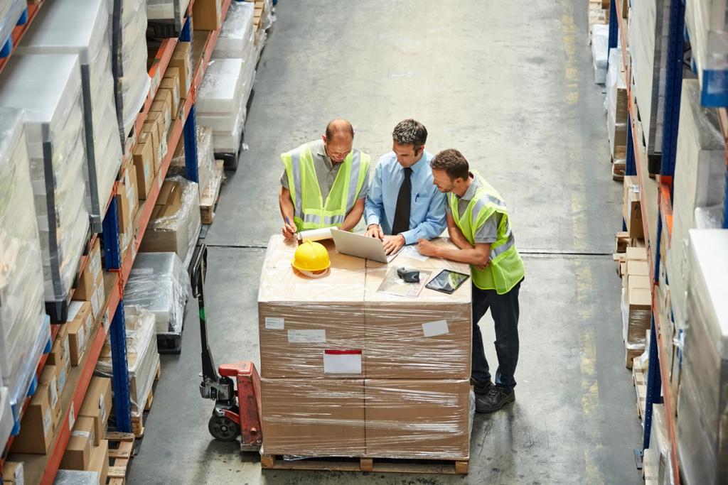 Three workers in hi-vis vests review a laptop on a stacked cardboard pallet in a warehouse aisle, hard hat nearby