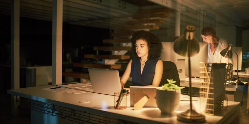 Woman works on a laptop at a desk in a dimly lit office at night, a colleague behind her.