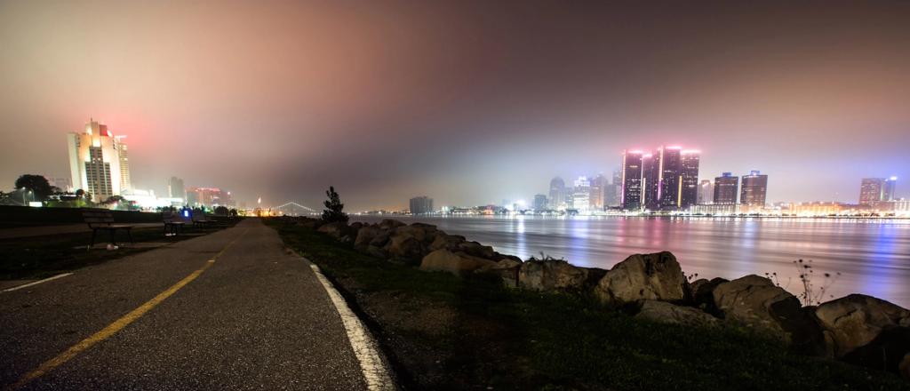 Nighttime waterfront with a lit city skyline reflected across the river and a foreground path