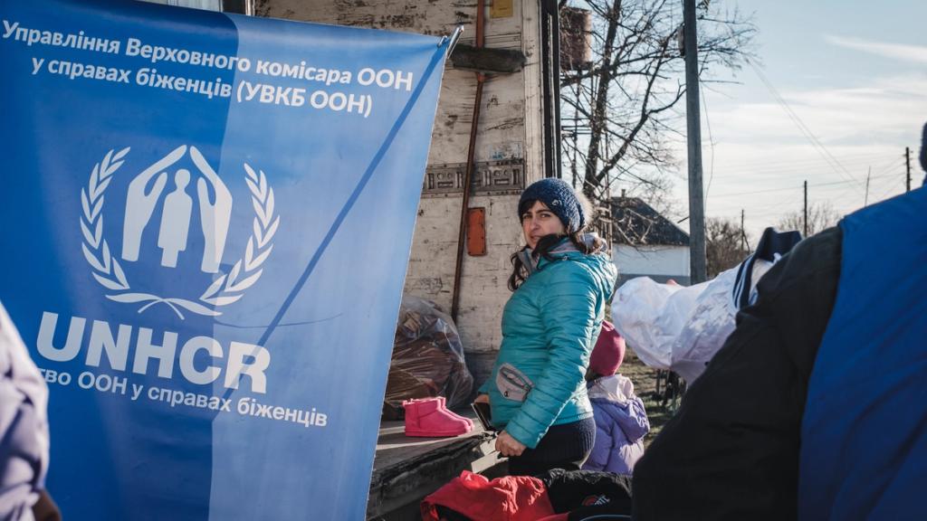 Woman by a UNHCR banner in Ukrainian beside a truck bed of donated clothing and aid