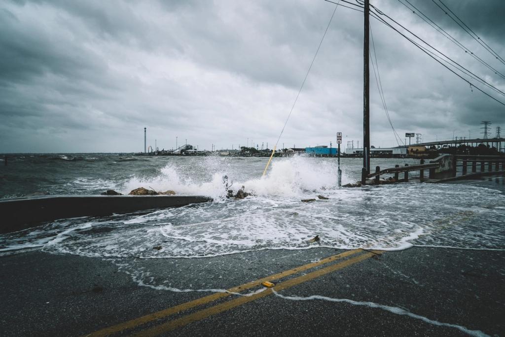 Storm-driven waves wash over a flooded coastal road beneath a dark overcast sky and power lines.