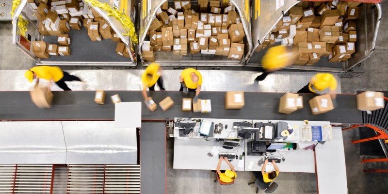 Aerial view of warehouse workers in yellow vests sorting boxes along conveyor belts and packing stations