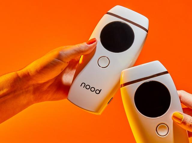 Two hands holding white Nood handheld IPL hair removal devices against an orange background