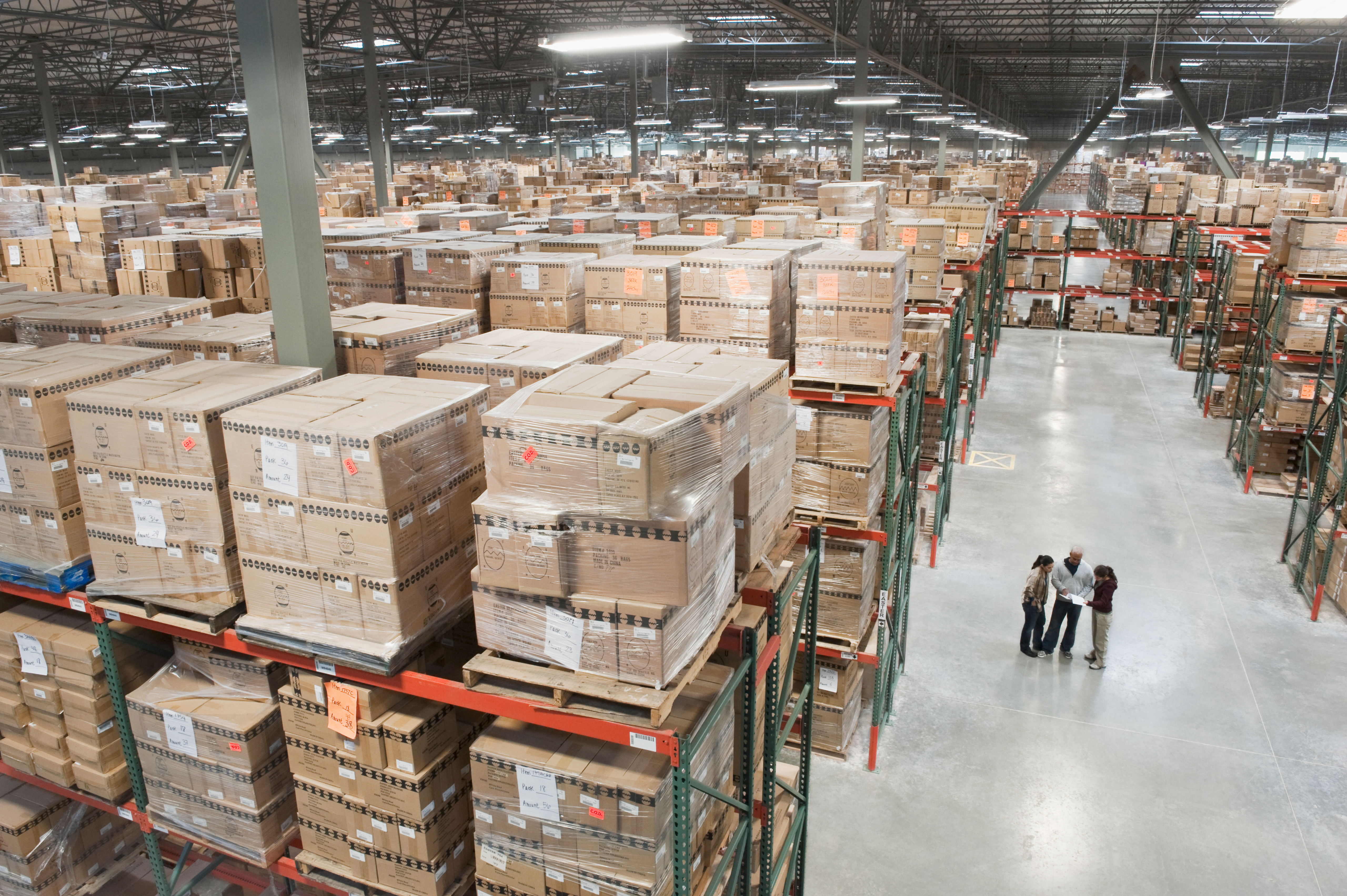 Workers inspect inventory in a large warehouse filled with cardboard boxes stacked on storage shelves.
