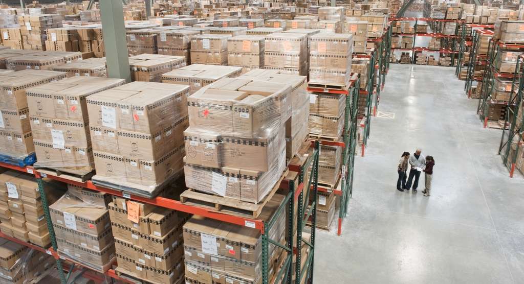 Workers inspect inventory in a large warehouse filled with cardboard boxes stacked on storage shelves.