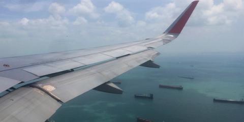 View from an airplane window of a red-tipped wing over teal water dotted with anchored cargo tankers