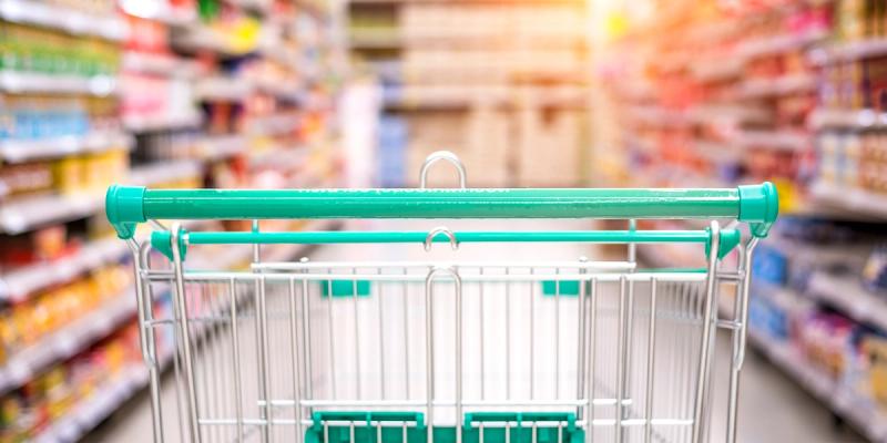 View pushing a teal shopping cart down a blurred, brightly stocked supermarket aisle.