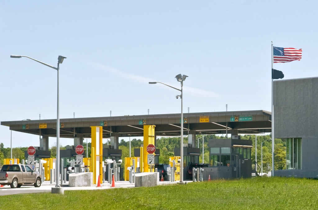 U.S. border crossing checkpoint with numbered inspection lanes, stop signs, and an American flag flying beside the station