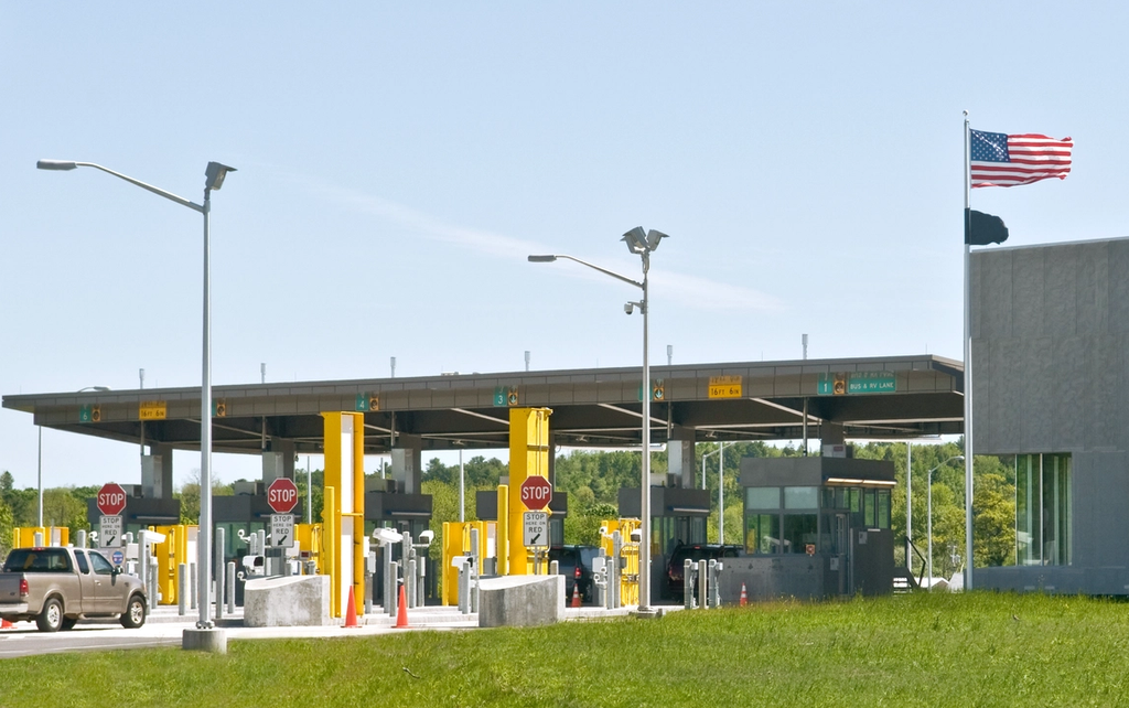 U.S. border crossing checkpoint with numbered inspection lanes, stop signs, and an American flag flying beside the station