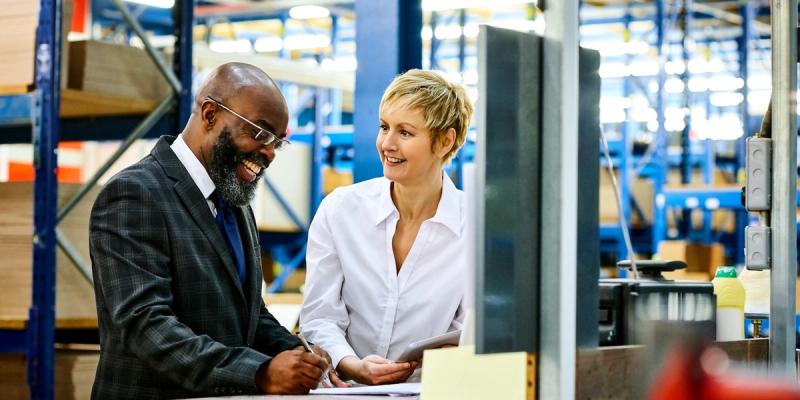 Smiling man in suit and woman in white shirt review paperwork together at a warehouse counter